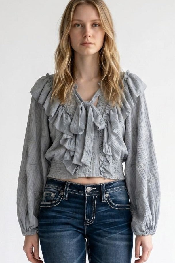 A woman wearing a light gray ruffled long-sleeve blouse with a tie-neck. Miss Me, blouse, top, love shack fancy, romantic, boutique